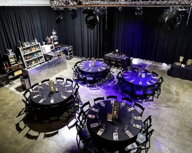 A spacious event room arranged with round tables covered in black tablecloths, a bar area in the background, and dim lighting from overhead fixtures.
