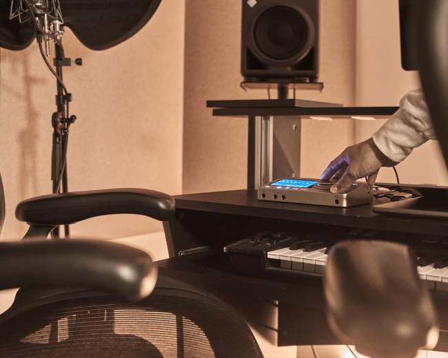 A hand is adjusting a device on a music production desk with a keyboard and studio monitors in a recording studio.