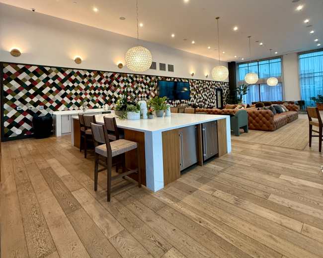 The image shows a modern common area featuring an open kitchen with a seating island, a patterned wall backdrop, and comfortable couches arranged for social gatherings.