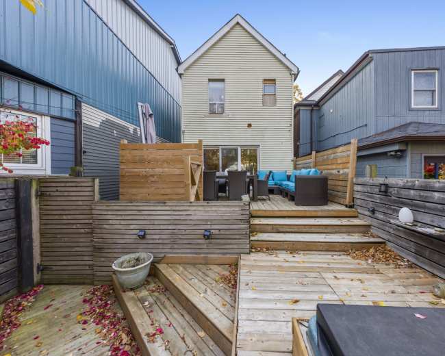 The image shows a backyard patio with wooden decking, featuring seating areas, a plant pot, and buildings in the background.