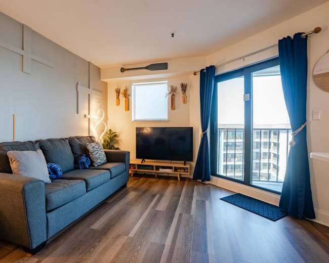 A modern living room features a gray sofa facing a television, with large windows providing natural light and access to a balcony.
