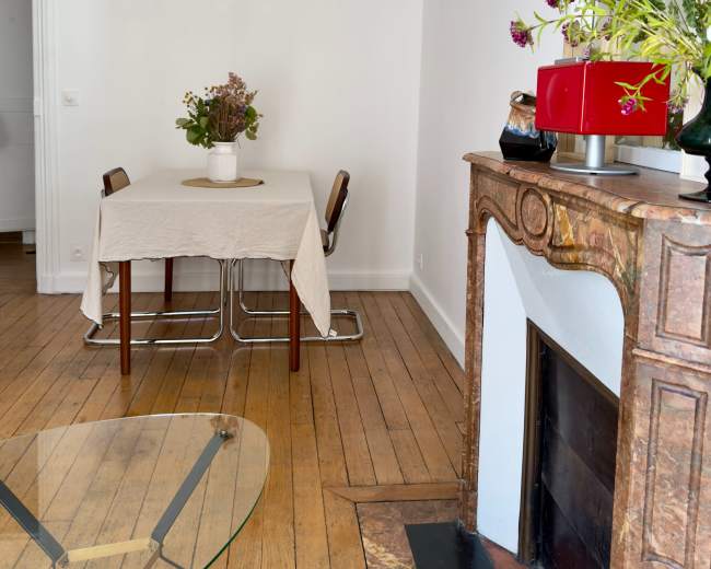 The image features a simple dining area with a table covered by a cloth, two chairs, a decorative mantel, and a vase of flowers on a nearby table.