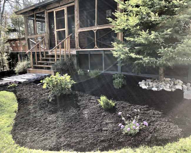 A screened porch with wooden steps is surrounded by freshly mulched garden beds featuring various plants and a small decorative rock arrangement.
