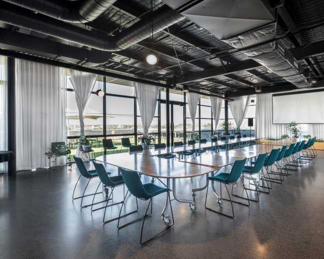 A large conference room features a long table with teal chairs arranged in a U-shape, surrounded by windows allowing natural light and equipped with a projector screen.