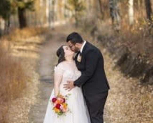 A couple stands on a leaf-covered path, sharing a kiss while the bride holds a bouquet of flowers.