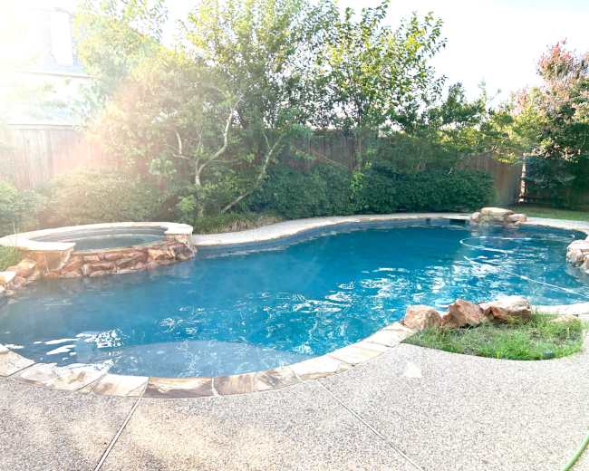 The image shows a landscaped backyard featuring a swimming pool with a stone border and a connected hot tub.