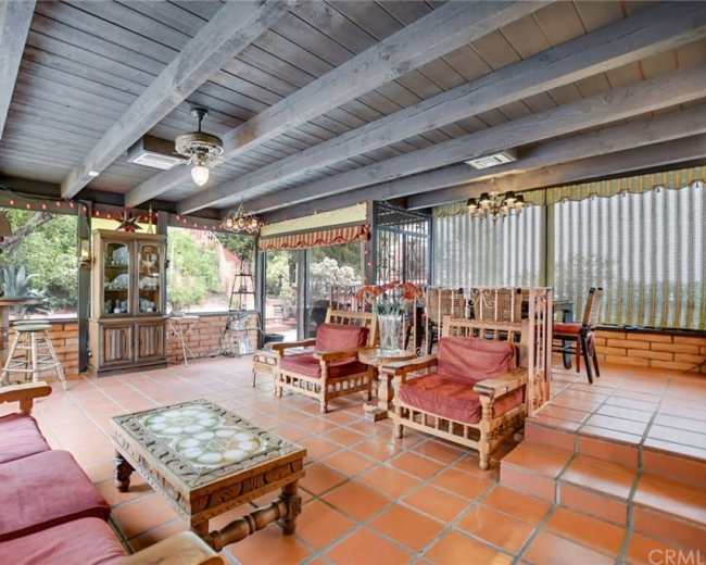 The image shows a spacious sunroom with terracotta tiles, wooden beams, and a mix of seating areas, including chairs and a coffee table.