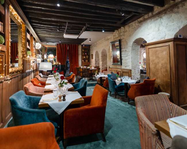 A restaurant interior features a mix of colorful seating, wooden beams, and softly lit tables arranged along a carpeted floor.