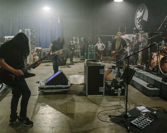 A band performs in a dimly lit rehearsal space filled with musical equipment and scattered crew members.