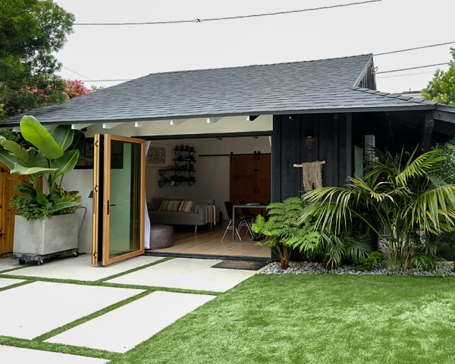 A small modern building with large sliding doors is surrounded by greenery and features a swimming pool nearby.