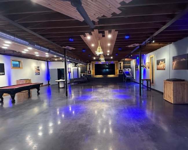 The image shows a spacious, modern entertainment room with polished concrete floors, blue lighting, and various wooden accents.