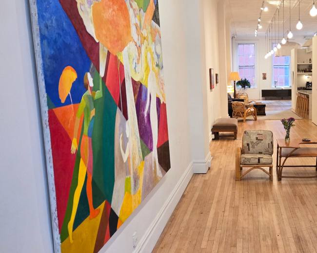 The image shows a spacious, well-lit art gallery with colorful artwork on the walls and seating areas in the center.