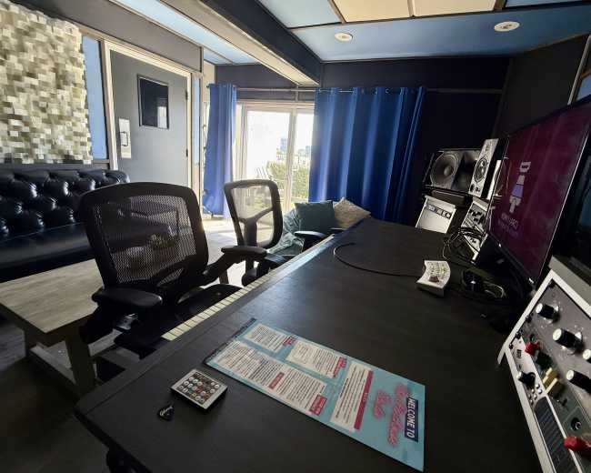 A modern music studio is set up with a desk featuring a keyboard and monitor, complemented by a comfortable seating area and blue curtains.