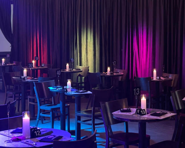 The image shows a dimly lit dining area with multiple tables, each set for guests and adorned with candles, against a backdrop of colorful illuminated curtains.