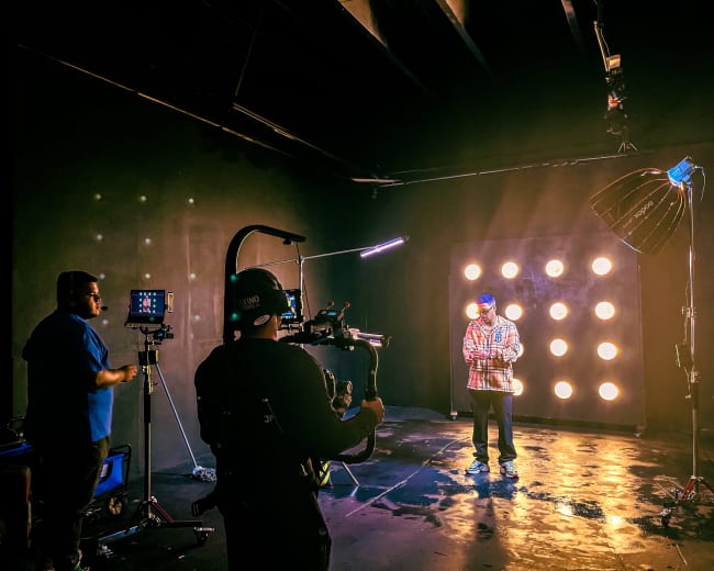 A person stands in a studio setting with a light fixture behind them, while two crew members operate filming equipment.