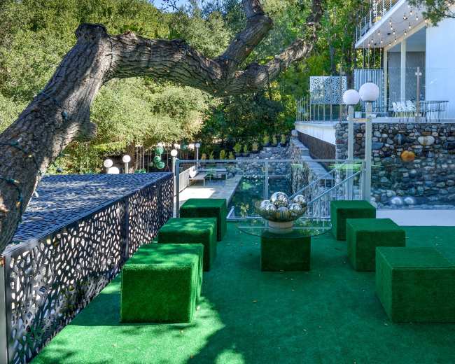 The image shows a modern outdoor patio area with green square seating, a decorative table featuring spherical ornaments, and a backdrop of trees and a stylish building.
