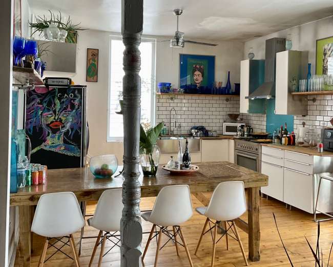 A modern kitchen features a wooden dining table surrounded by white chairs, with colorful artwork and plants on the walls and shelves.