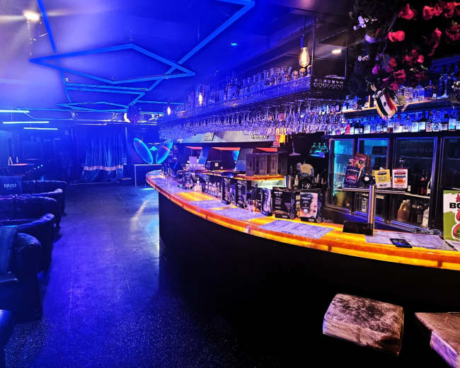 The image shows a dimly lit bar featuring a curved illuminated counter, rows of hanging glassware, and various seating options arranged around the space.