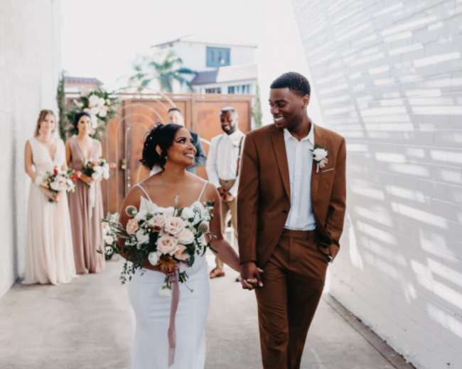 A bride and groom walk hand in hand down a pathway, accompanied by their wedding party in the background.