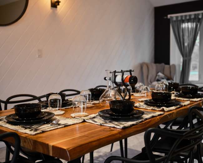 A wooden dining table is set with black dishes, glassware, and utensils, under soft lighting in a modern room.