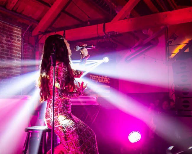 A singer performs on stage in a dimly lit venue with colorful lights and a crowd in the background.