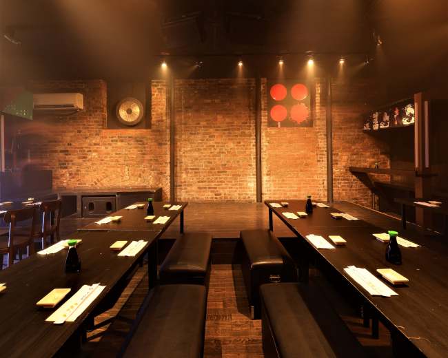 A dimly lit restaurant with brick walls features long, dark wooden tables and benches arranged for dining.