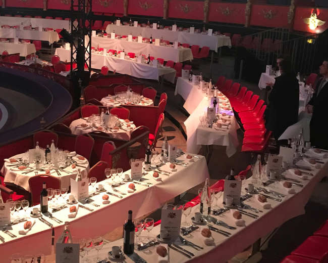 The image shows a circular banquet hall set with numerous elegantly arranged dining tables surrounded by red chairs.