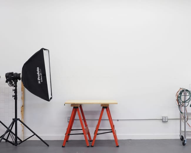 The image shows a simple studio setup featuring a black softbox light on a stand, a wooden work table on orange sawhorses, and a metal rack with coiled cords against a white wall.