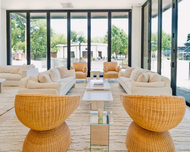 A modern living room features beige sofas and rattan chairs arranged around a glass coffee table, with large windows providing a view of the outdoors.