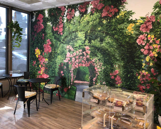 The image shows a café interior featuring a large floral mural on one wall, with a seating area that includes small tables and chairs, and a display case for pastries.