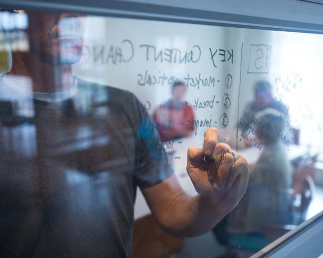 A person writes on a glass board while others sit at a table in the background.