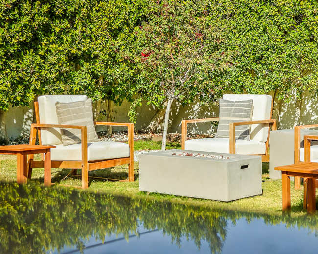 A set of four wooden lounge chairs with cushions and two small tables is arranged on a grassy area surrounded by shrubs.