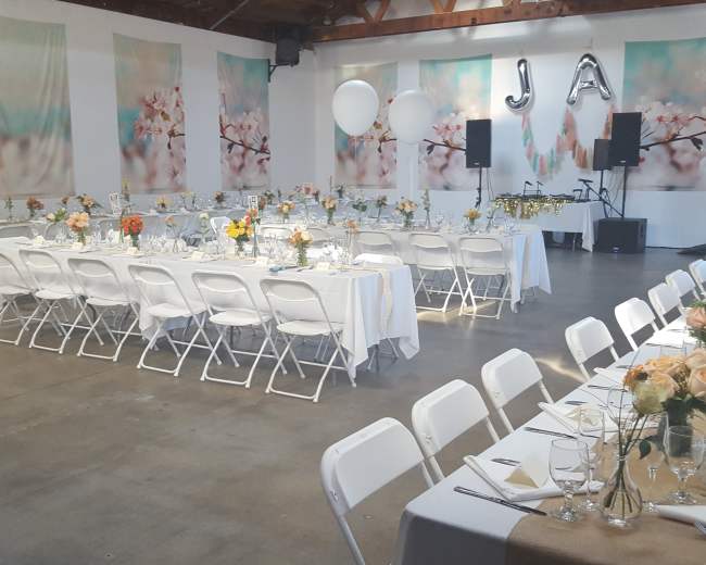 A large indoor event space is set up for a celebration, featuring long tables adorned with floral centerpieces and a backdrop of soft, cherry blossom-themed decorations.