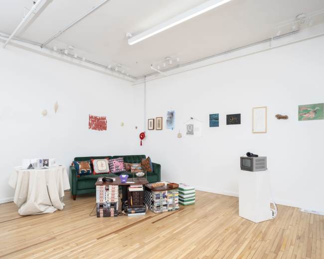 The image shows a modern art gallery featuring a green sofa, a table covered with a cloth, and various artworks displayed on the walls.