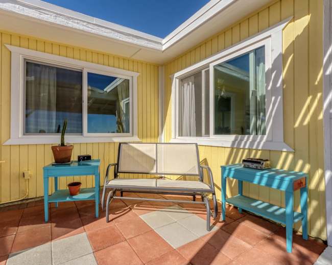 A small patio area features a beige bench flanked by two turquoise side tables, with potted plants and large windows set into a yellow wall.