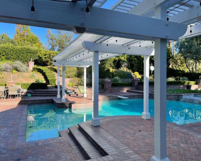 The image shows a spacious pool area surrounded by brick paving, with a covered patio and landscaped greenery in the background.