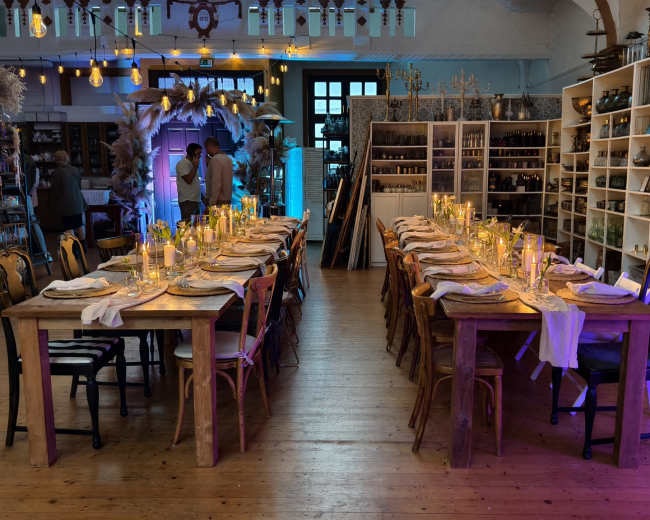 The image shows a spacious dining area with two long wooden tables set for a meal, adorned with candles and tableware, surrounded by shelves displaying various decorative items.