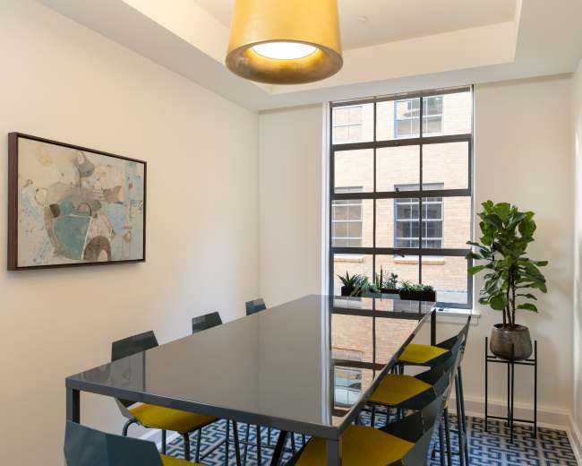 A modern conference room features a long glass table surrounded by colorful chairs, a large window with potted plants, and an abstract painting on the wall.