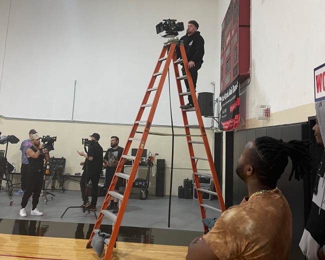 A man stands on a ladder, operating a camera in a gym while a group of people watch from below.