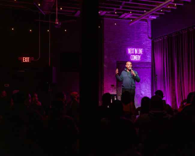 A comedian stands on stage in a dimly lit venue, delivering a performance to an audience seated in front of a curtain with purple lighting.