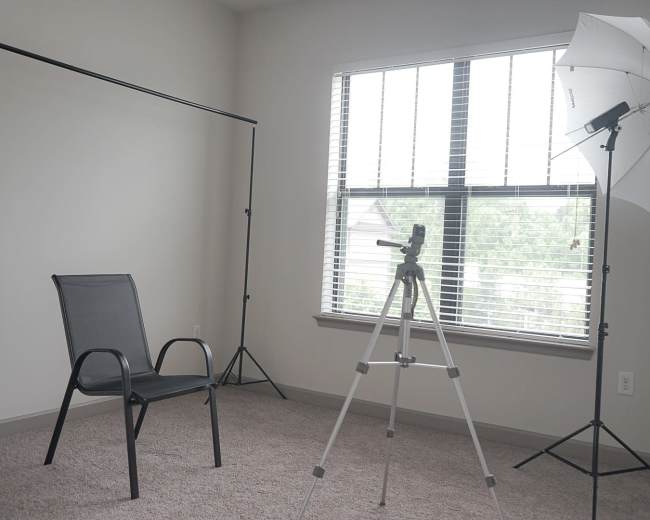 The image shows a bare room with a chair, a light stand with an umbrella, and a tripod positioned near a window with blinds.