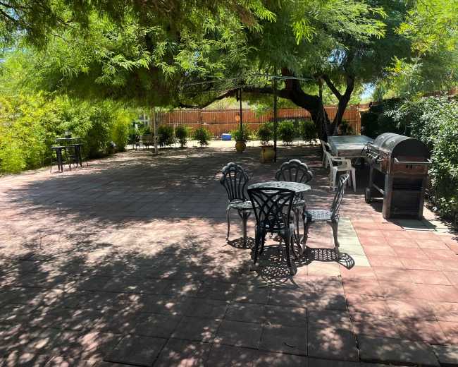 The image shows a spacious outdoor patio area with tiled flooring, shaded by trees, and equipped with metal furniture and a barbecue grill.