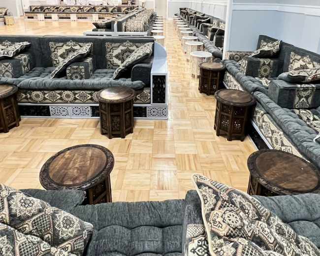 The image shows a spacious lounge area furnished with patterned couches and small wooden tables arranged around a wooden floor.
