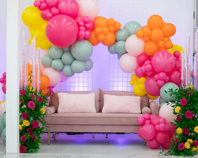 A sofa is positioned under a large arch of colorful balloons, surrounded by floral arrangements.