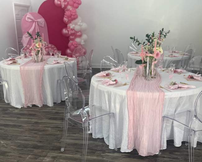 The image shows two elegantly set round tables with white tablecloths and pink decorations, including table runners and centerpieces, in a brightly lit venue.