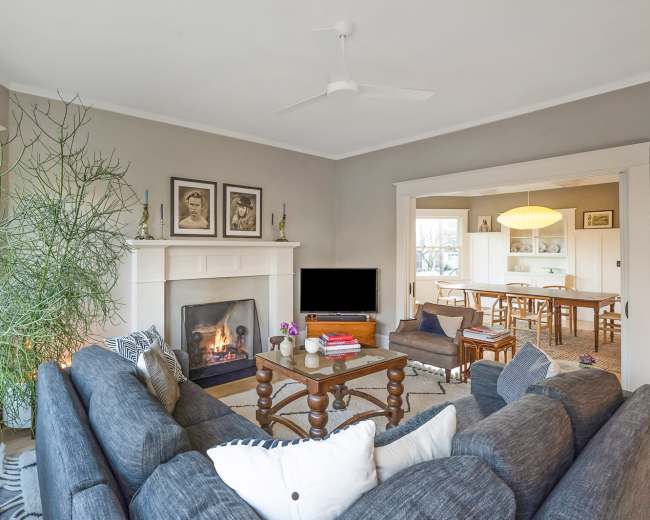 The living room features a gray sofa facing a fireplace, with a large plant beside it and an open view into a dining area.