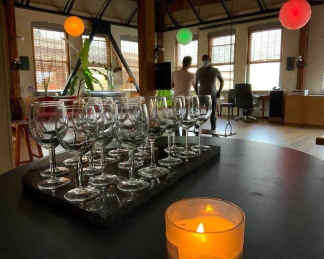 The image shows a table with a row of wine glasses in the foreground, while two people converse in a well-lit room decorated with hanging lanterns and potted plants.