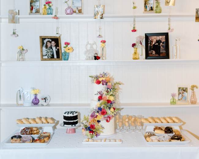 A decorated table with a cake and various pastries is set in front of shelves displaying framed photos and hanging floral arrangements.