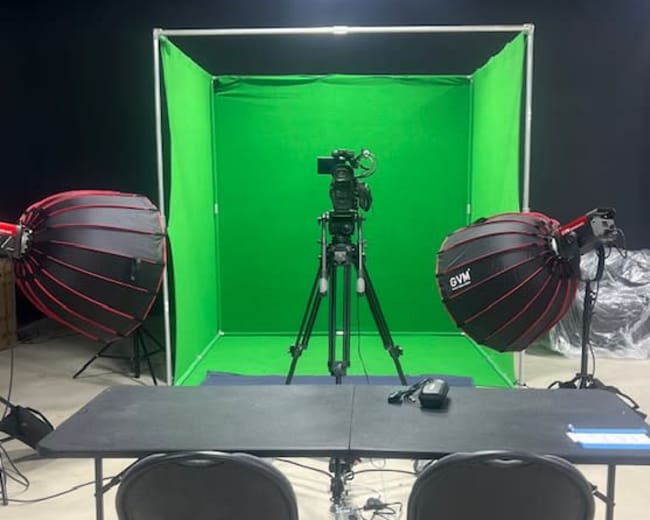 A green screen setup featuring a camera on a tripod and two large light sources positioned on either side of a black table and chairs.