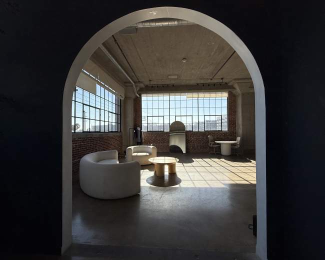 The image shows a spacious, industrial-style room with large windows, featuring a circular table and two white chairs.